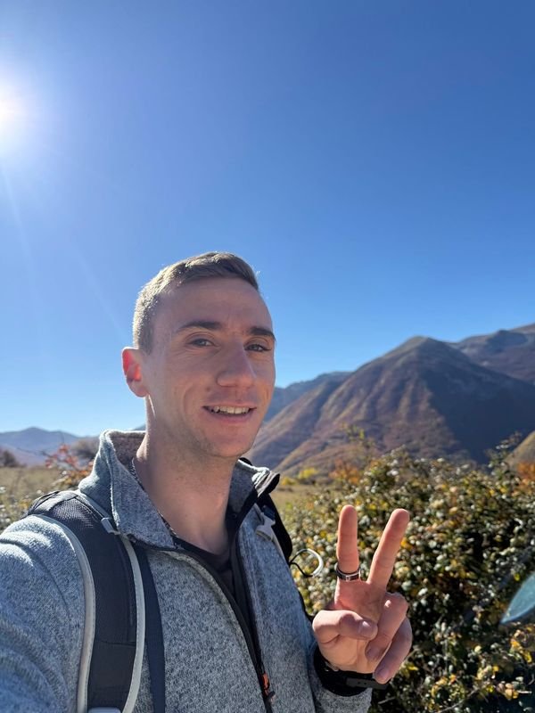 Giulio sorride e fa il segno di pace, con lo zaino in spalla, davanti a montagne in autunno